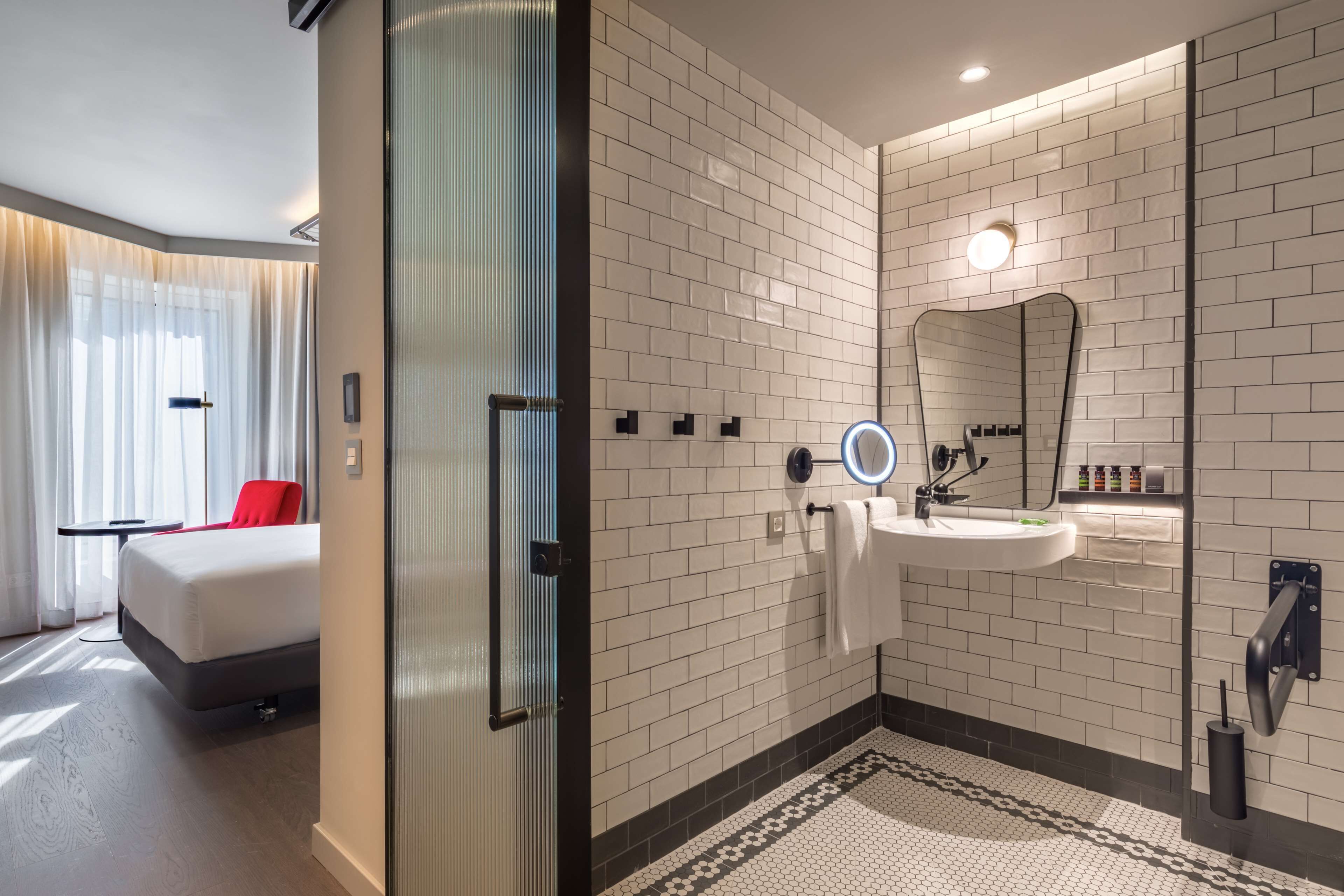 Une salle de bain d’hôtel moderne avec des murs recouverts de carreaux blancs, un grand miroir, un lavabo et un sol à motifs.