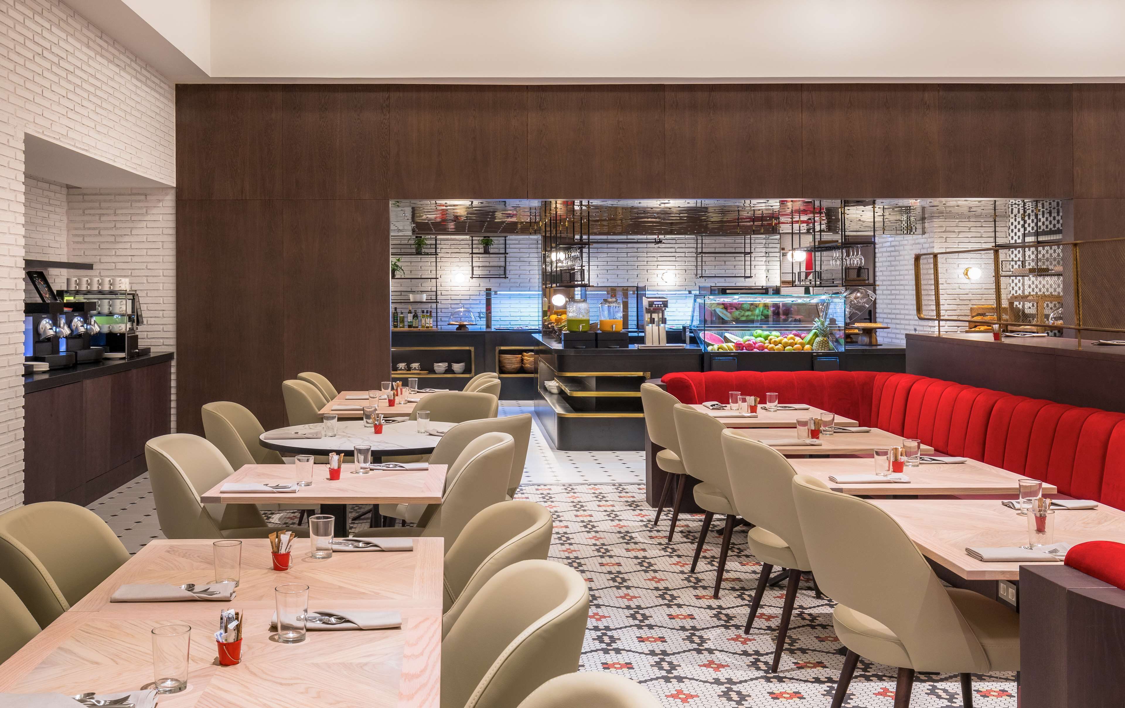 Un interno ristorante moderno con tavoli rotondi, sedie beige, una panca rossa e un angolo caffetteria.