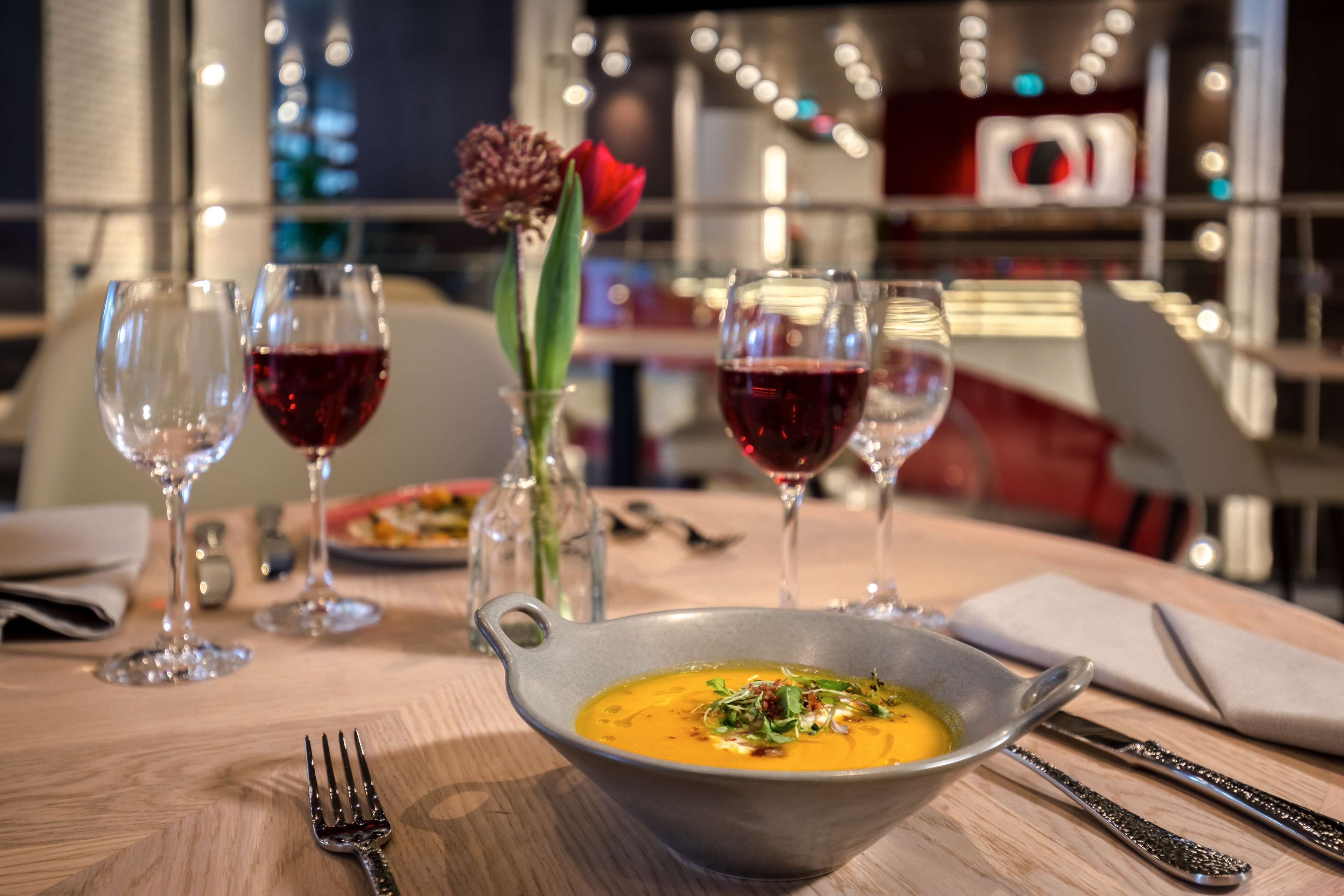A dining table with a bowl of soup, two wine glasses, a fork, and a rose in a vase.