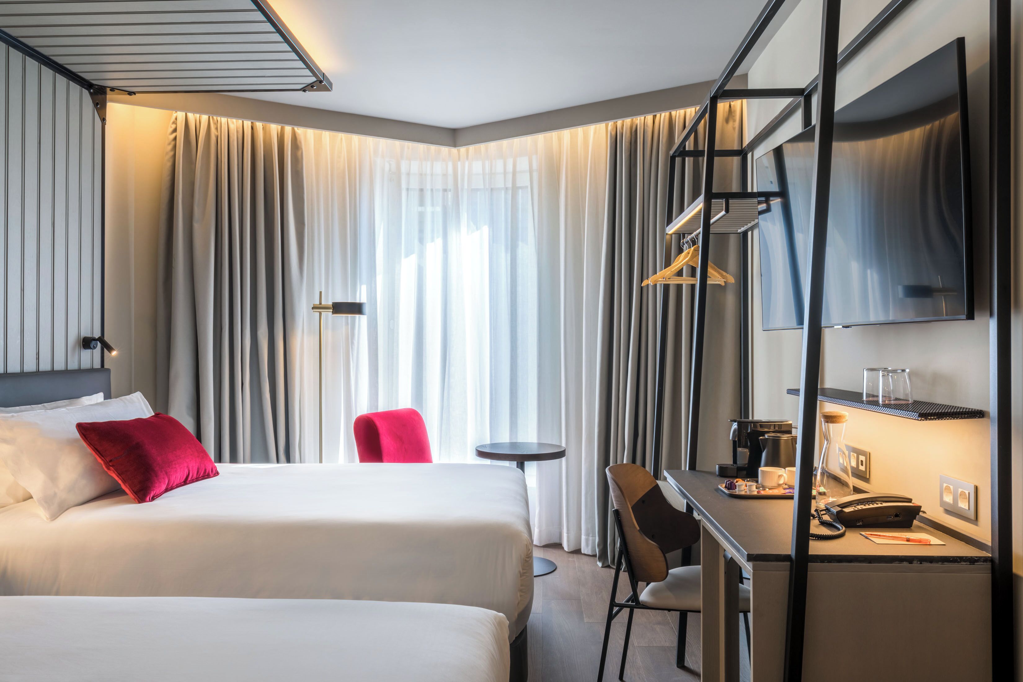 Habitación de hotel con dos camas, un escritorio con silla, una mesita pequeña, televisor y ventana con cortinas.