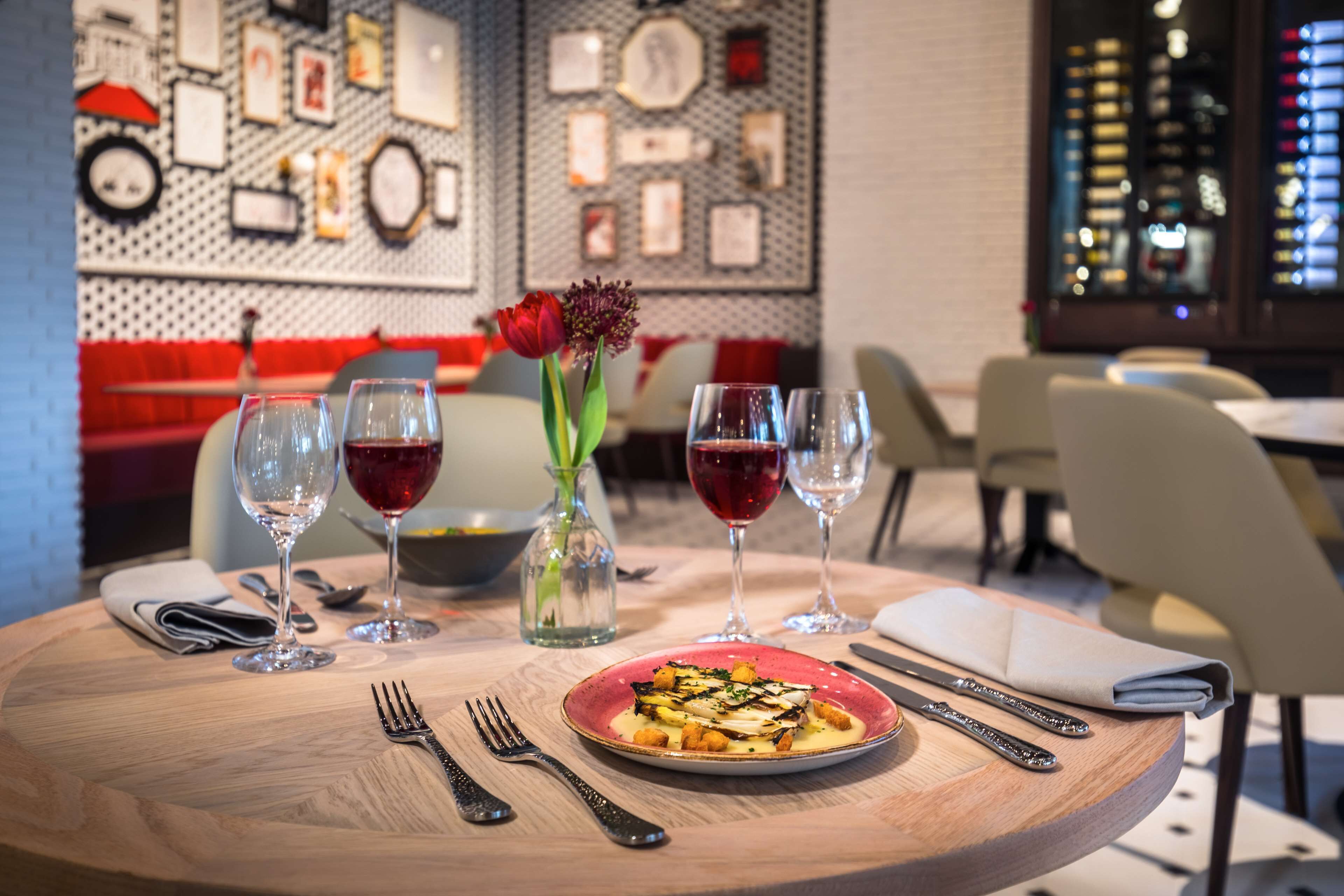 A dining table with a plate of food, wine glasses, cutlery, and a vase with a flower.