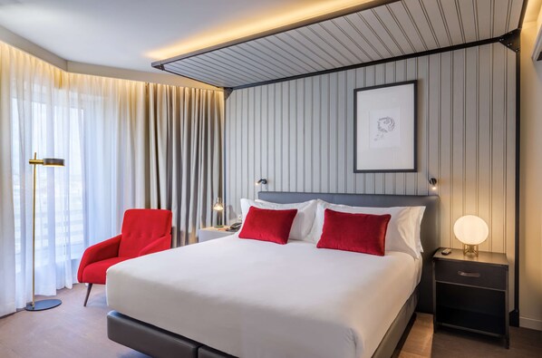 A modern hotel room with a large bed, red armchair, bedside table with lamp, and a framed picture on the wall. - Canopy by Hilton Madrid Castellana (Madrid)
