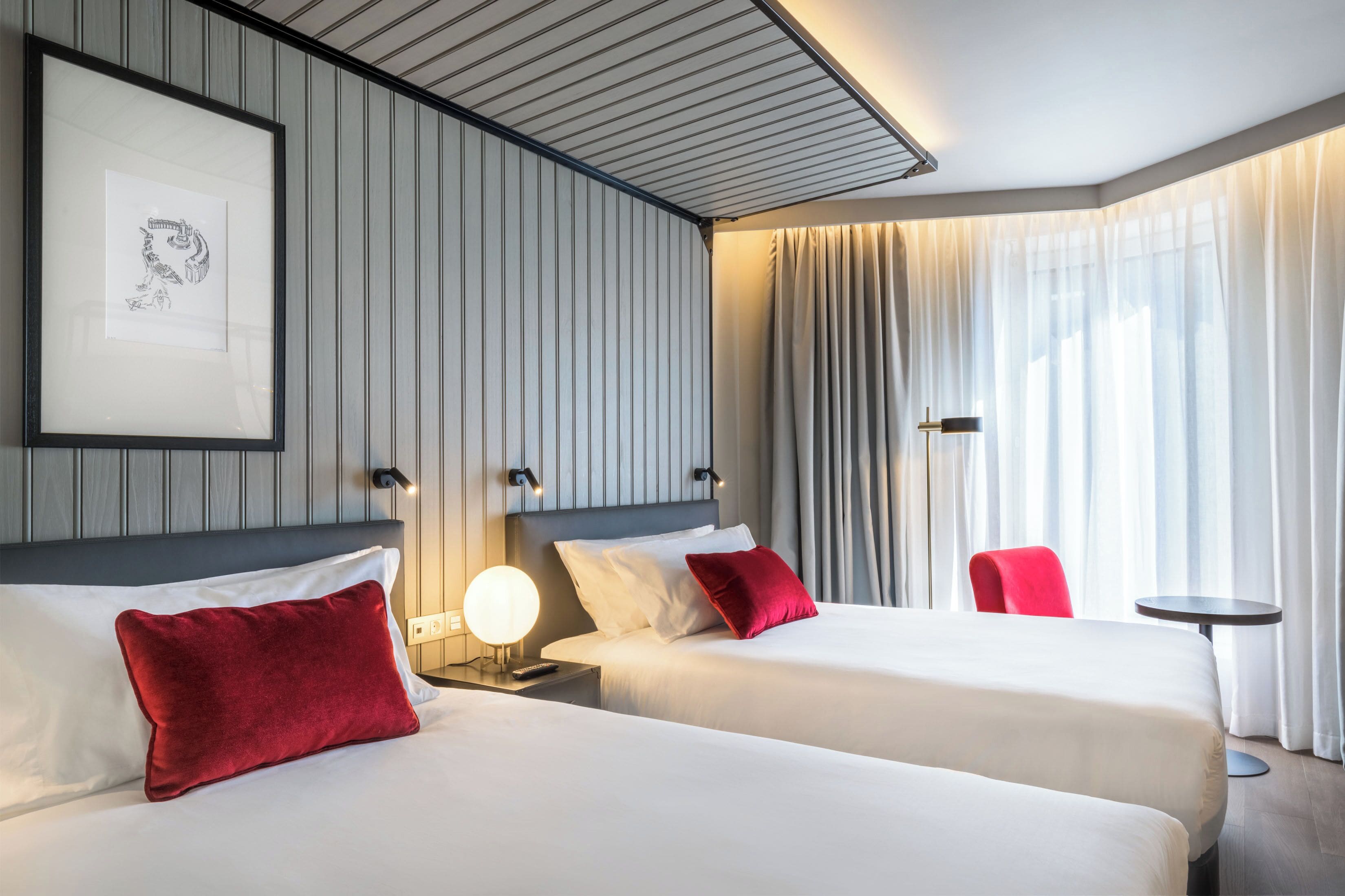 Habitación de hotel con dos camas, un cuadro en la pared, una mesita de noche con lámpara, una silla roja y una pequeña mesa redonda.