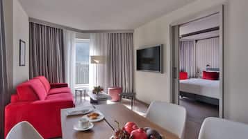 Quarto de hotel com um sofá vermelho, uma mesa de jantar com frutas, uma televisão e uma varanda com cortinas.