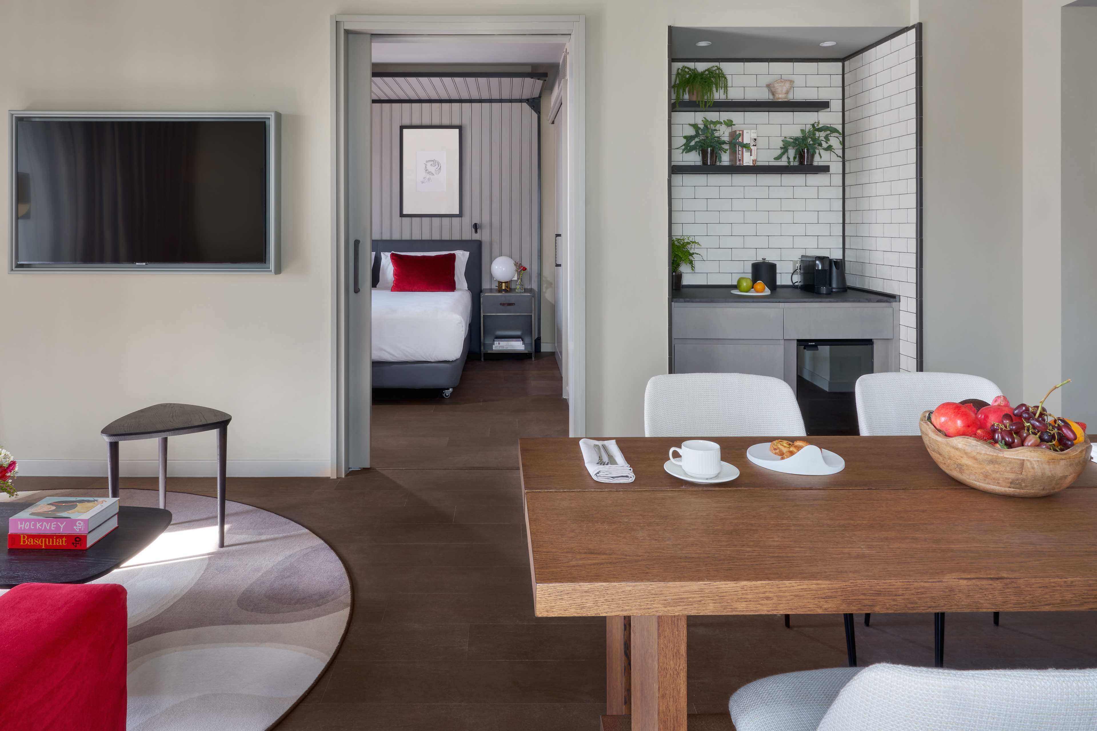Una habitación de hotel moderna con cama, televisor, zona de comedor con mesa y sillas, y cocina con estantes y electrodomésticos.