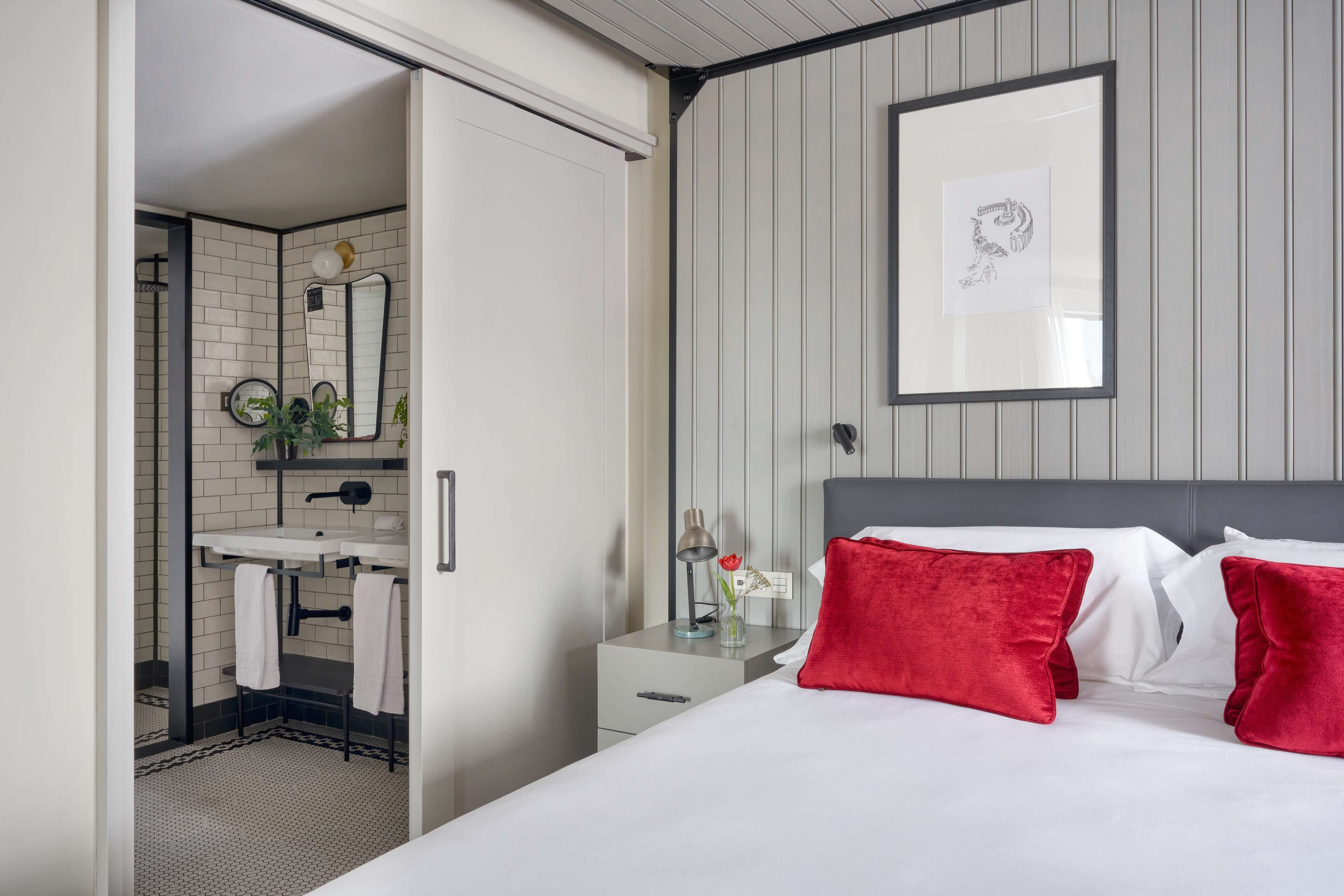 Une chambre d’hôtel avec un lit, une table de chevet, une commode, un miroir, un tableau encadré au mur et une salle de bain visible à travers une porte ouverte.