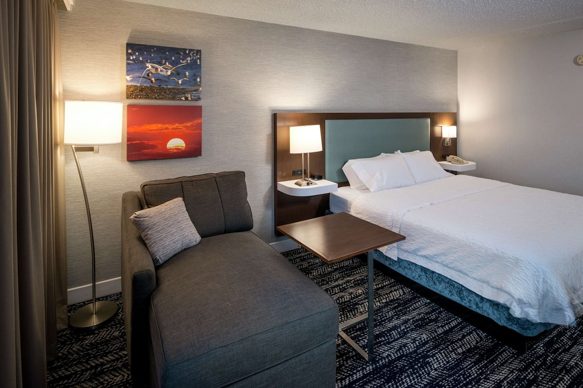 A hotel room with a bed, a sofa, a nightstand, a lamp, and two wall art pieces.