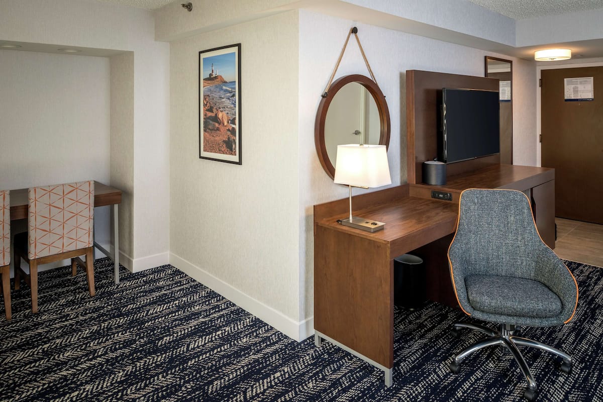 A hotel room with a wooden desk, a grey office chair, a wall-mounted TV, a desk lamp, and a framed picture of a lighthouse.