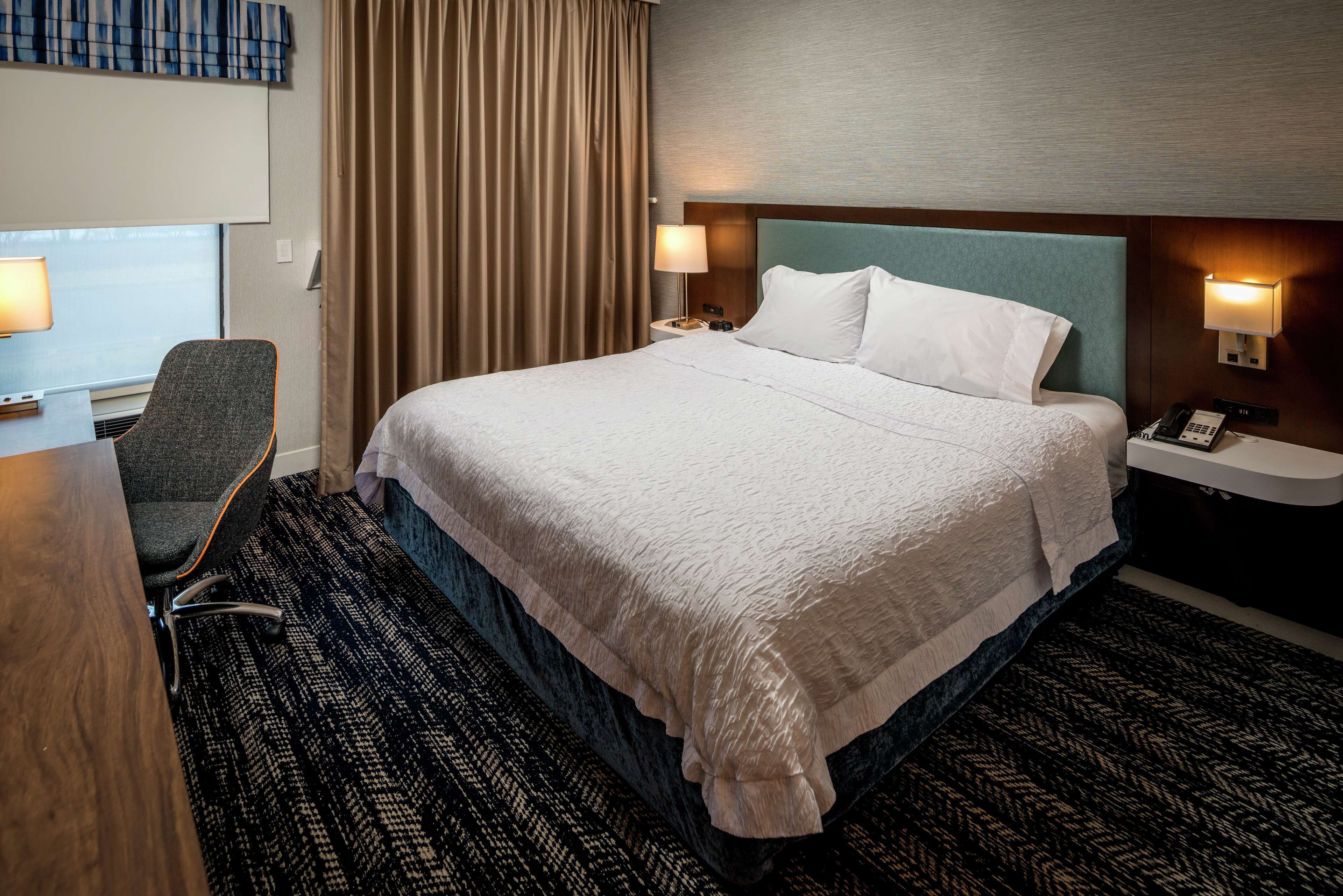 Une chambre d’hôtel avec un grand lit, un bureau avec une chaise, une lampe de bureau, un téléphone et un luminaire fixé au mur.