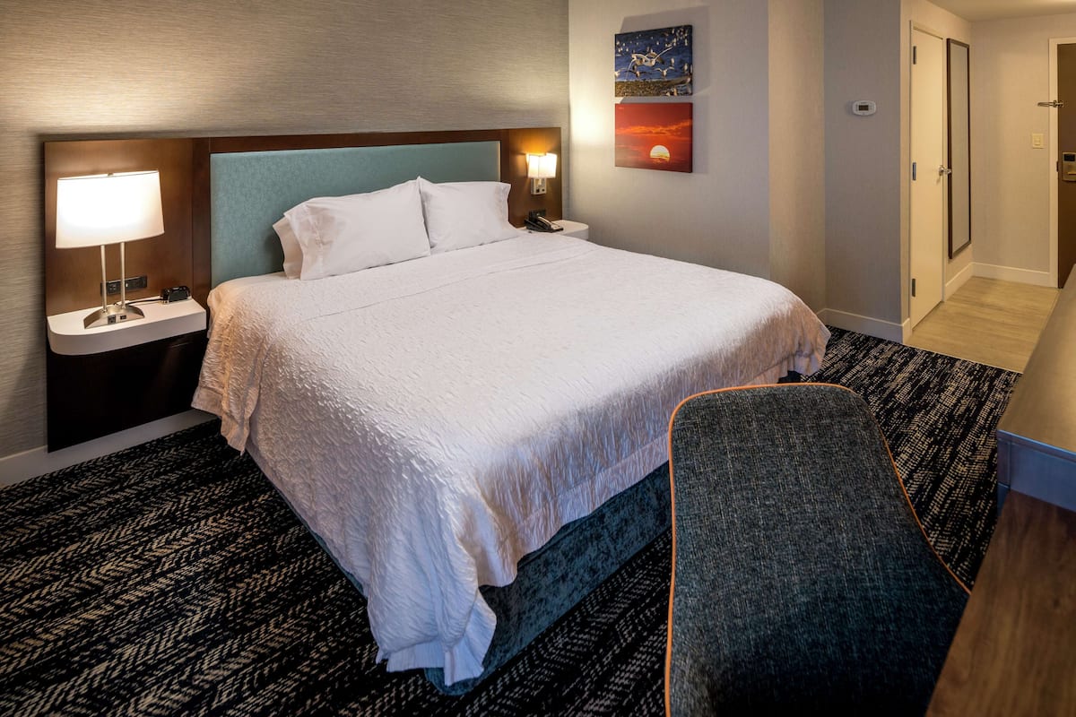 A hotel room with a large bed, a bedside table with a lamp, a chair, and a painting on the wall.