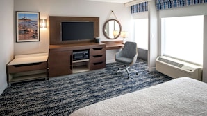 A hotel room with a bed, a desk with a flat-screen TV, a chair, a large window with curtains, and a wall-mounted mirror. - Hampton Inn Long Island / Commack (Commack)