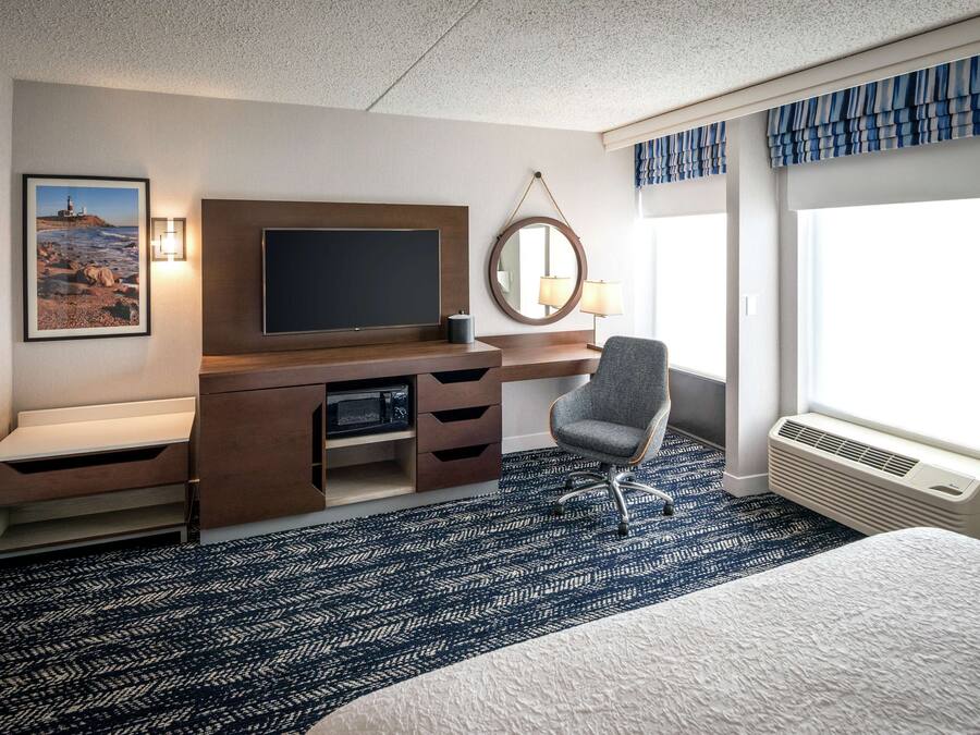 Habitación de hotel con cama, escritorio con televisor de pantalla plana, una silla, ventana grande con cortinas y espejo instalado en la pared.