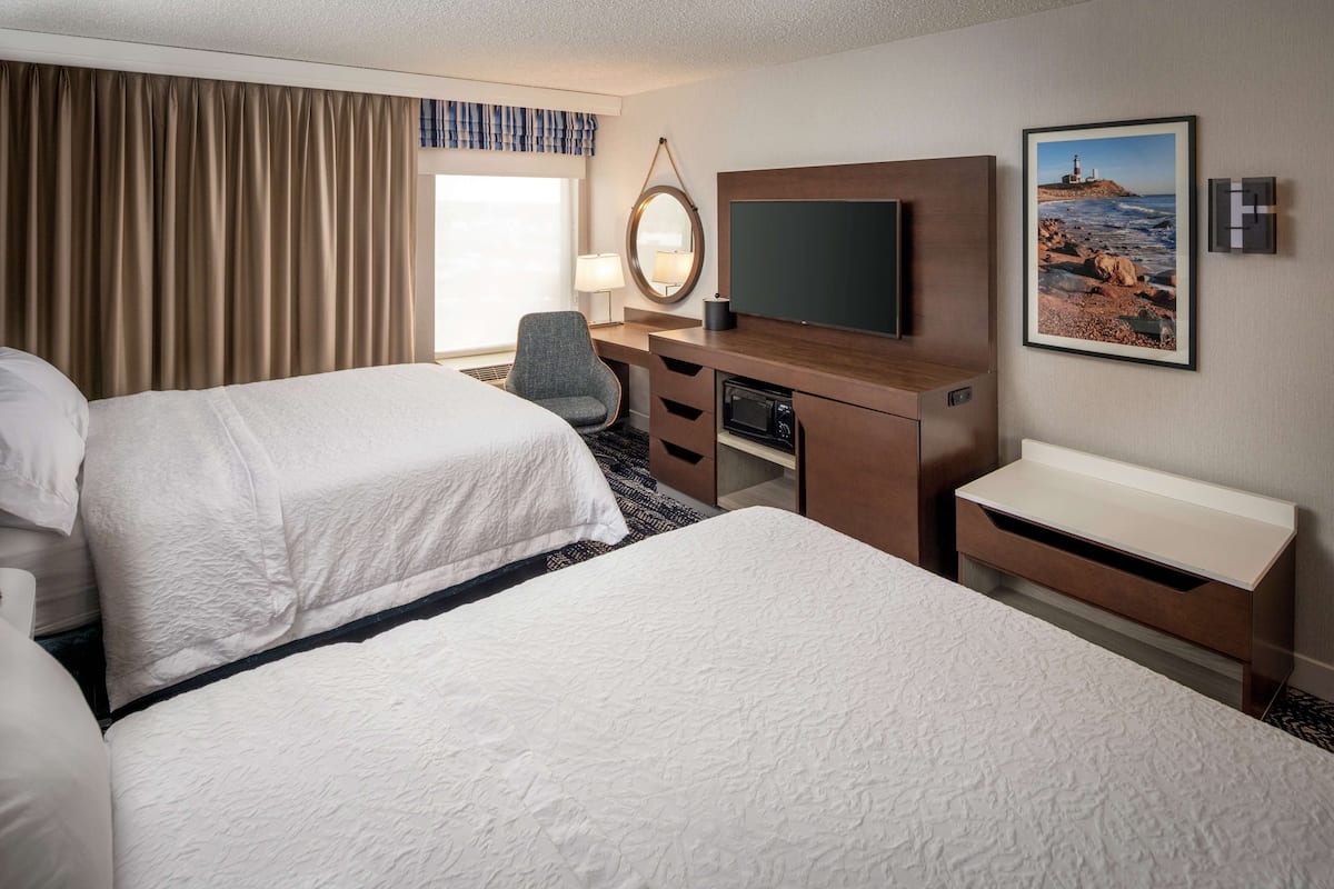 A hotel room with two beds, a desk with a flat-screen TV, a chair, a desk lamp, and a framed picture on the wall.