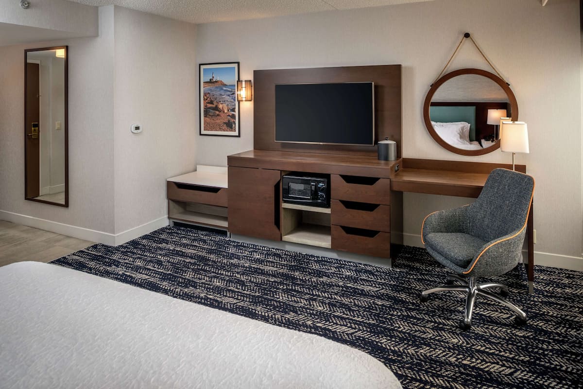 A hotel room with a bed, a desk with a flat-screen TV, a chair, a round mirror, and a wall-mounted lamp.