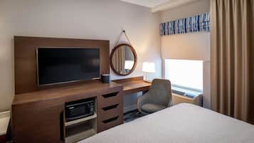 A hotel room with a large bed, a flat-screen TV, a desk with a microwave, a chair, and a window with curtains.