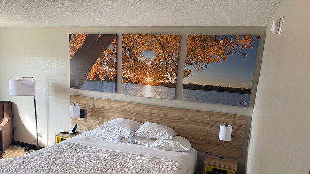 A hotel room with a large bed, wooden headboard, bedside tables, and wall-mounted artwork depicting autumn leaves and a sunset over water. - Days Inn by Wyndham Muncie -Ball State University (Muncie)