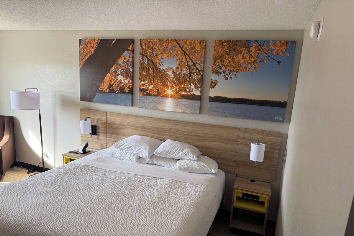 A hotel room with a large bed, wooden headboard, bedside tables, and wall-mounted artwork depicting autumn leaves and a sunset over water.