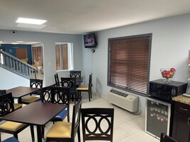Un comedor con mesas y sillas, un televisor montado en la pared, una ventana con persianas y un refrigerador con un frutero encima.