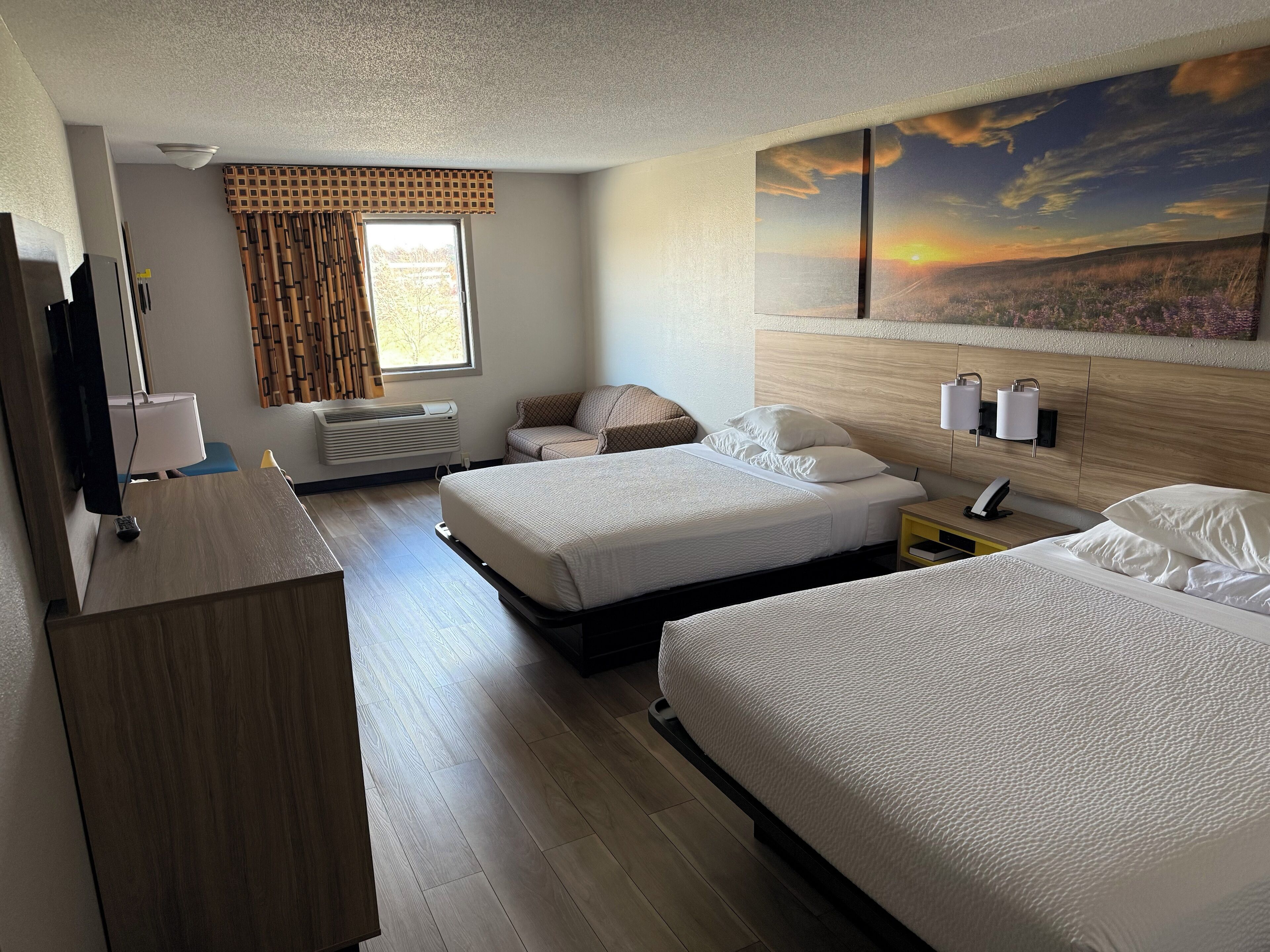 A hotel room with two beds, a desk, a TV, and a painting of a sunset.