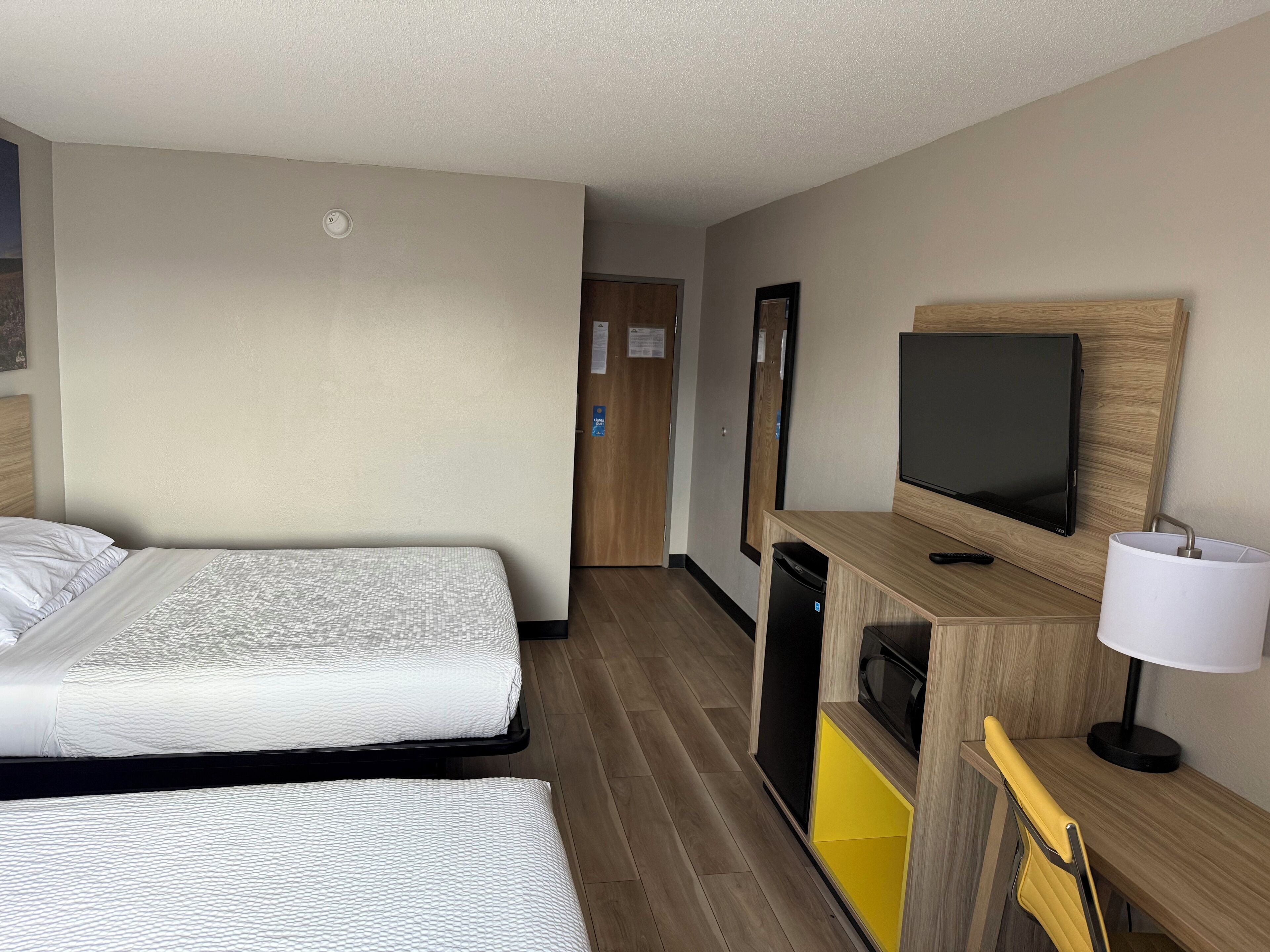 A hotel room with two beds, a TV, a desk with a lamp, and a small kitchenette.