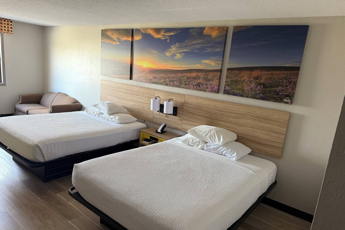 A hotel room with two beds, a sofa, and a wall-mounted artwork depicting a sunset.
