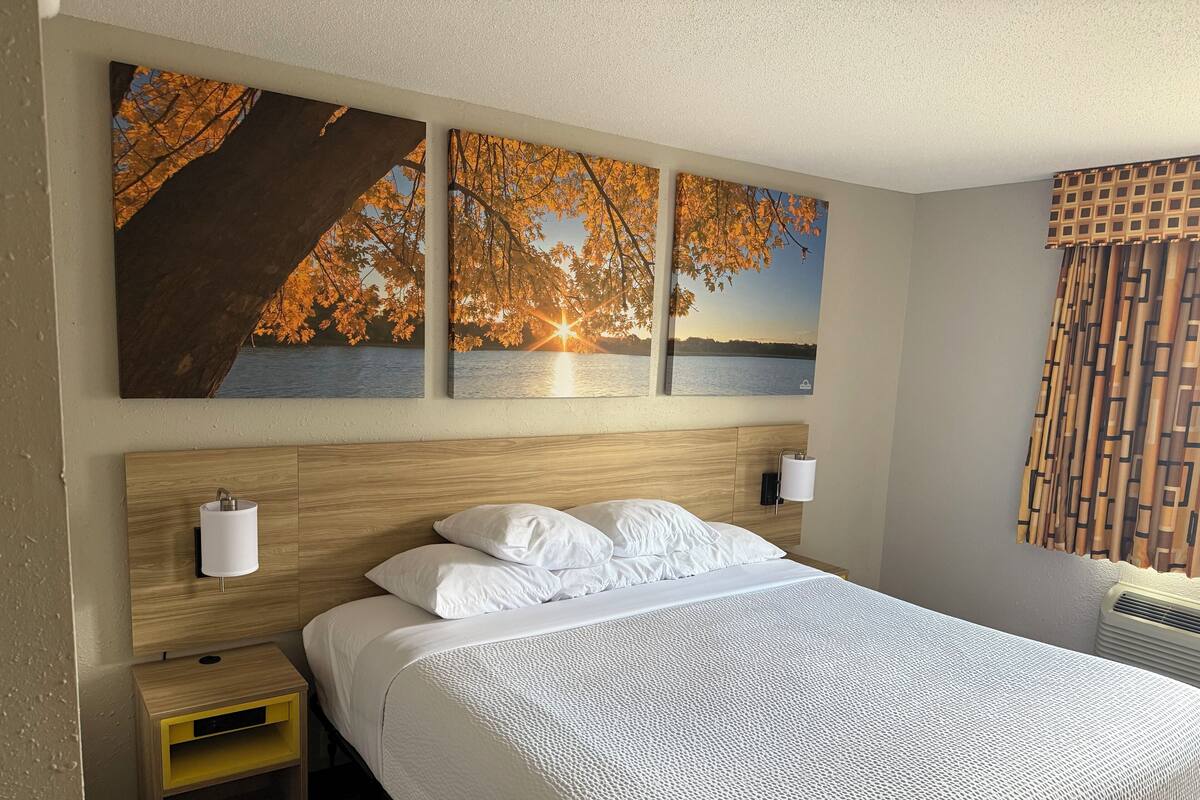 A hotel room with a large bed, two bedside lamps, a nightstand, and three wall-mounted paintings of a sunset over water.