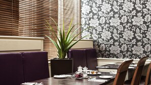 A restaurant interior with a table set for two, purple booth seating, a potted plant, and a floral wallpaper pattern.