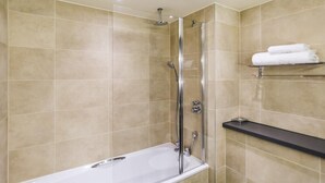 A modern bathroom with a glass-enclosed shower, a white bathtub, beige tiled walls, and a black shelf with towels.