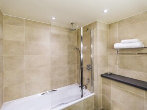 Une salle de bain moderne avec une douche à parois de verre, une baignoire blanche, des carreaux de couleur beige et une étagère noire où sont posées des serviettes.