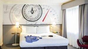 A hotel room with a bed, two bedside tables, a clock on the wall, and a window with curtains.