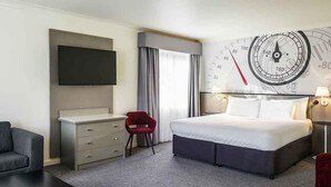 A hotel room with a bed, a desk, a TV, a clock, and a red chair.