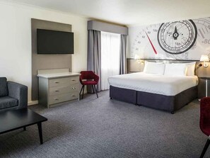 Ein Hotelzimmer mit einem Bett, einem Schreibtisch, einem Fernseher, einer Uhr und einem roten Sessel.