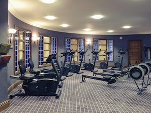A gym with various exercise machines, including treadmills and elliptical trainers, arranged in a spacious room with patterned flooring and large windows.