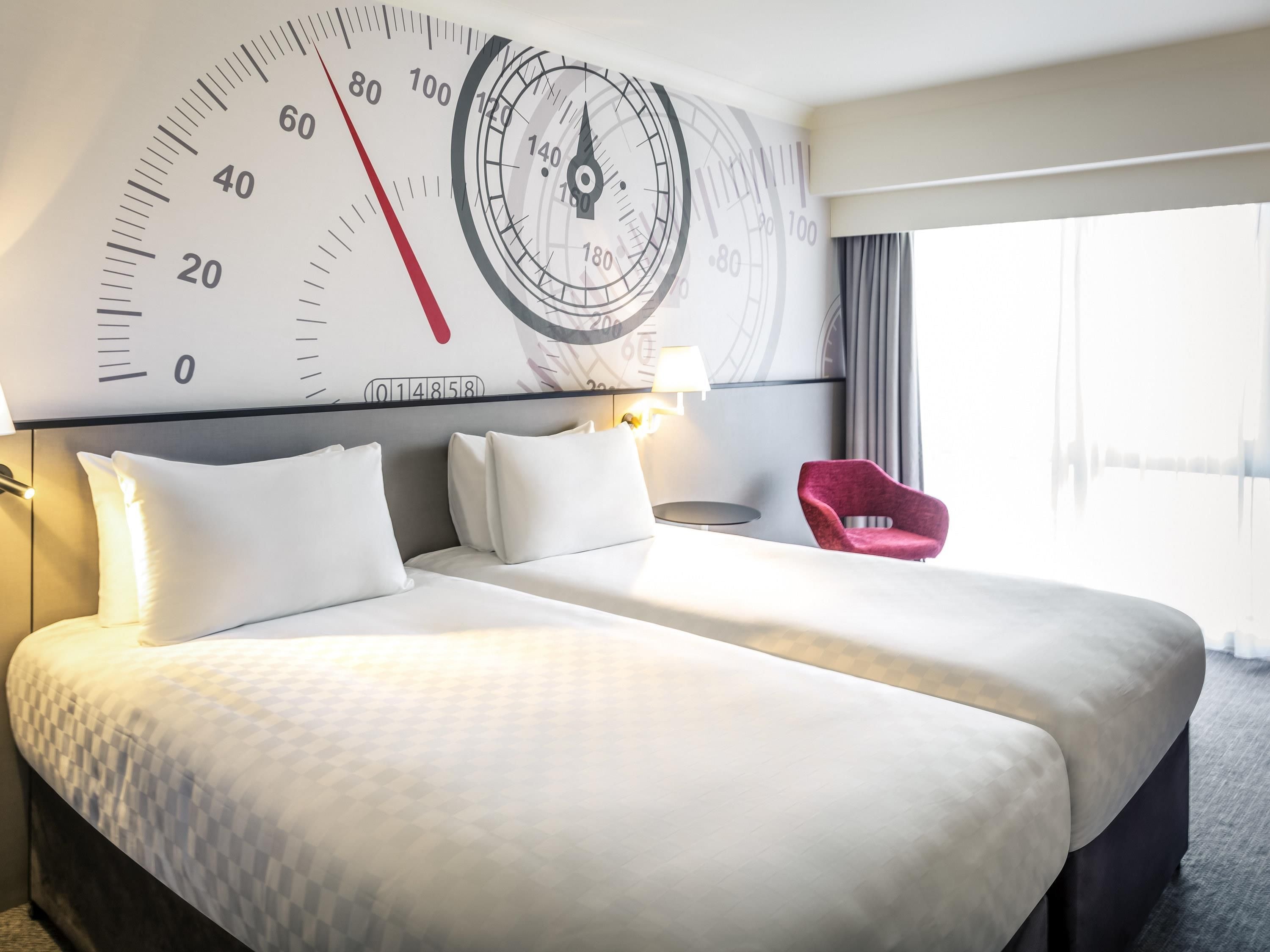 Habitación de hotel con un reloj de pared grande, una cama con ropa de cama blanca, una silla roja y una lámpara.