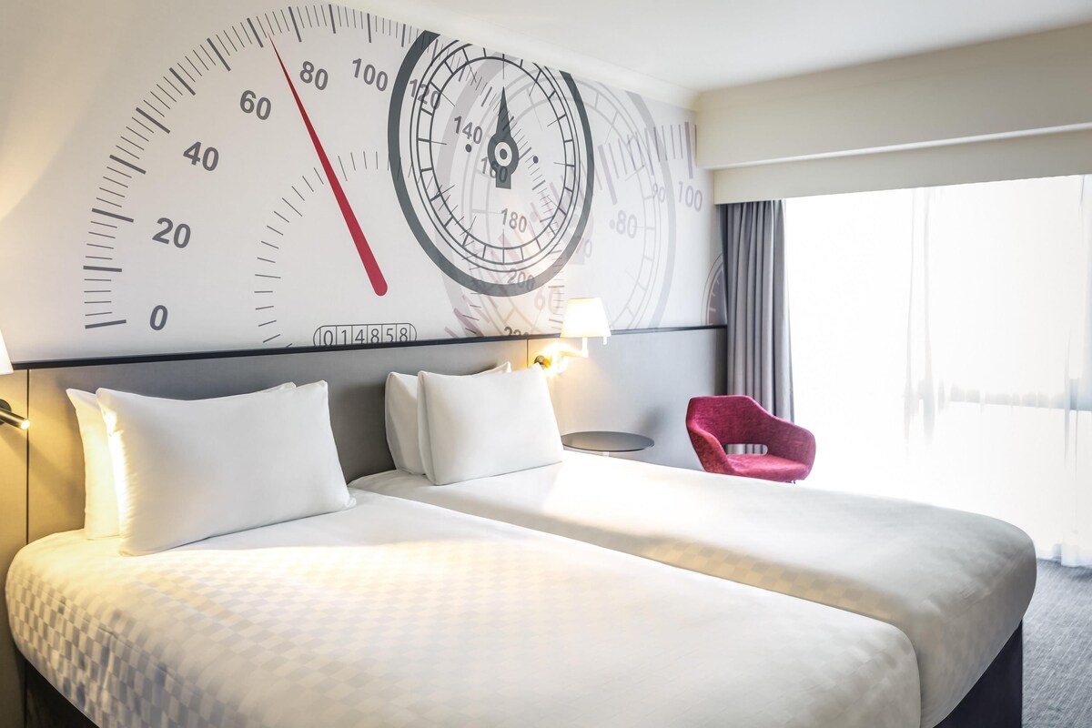Een hotelkamer met een grote wandklok, een bed met wit beddengoed, een rode stoel en een lamp.