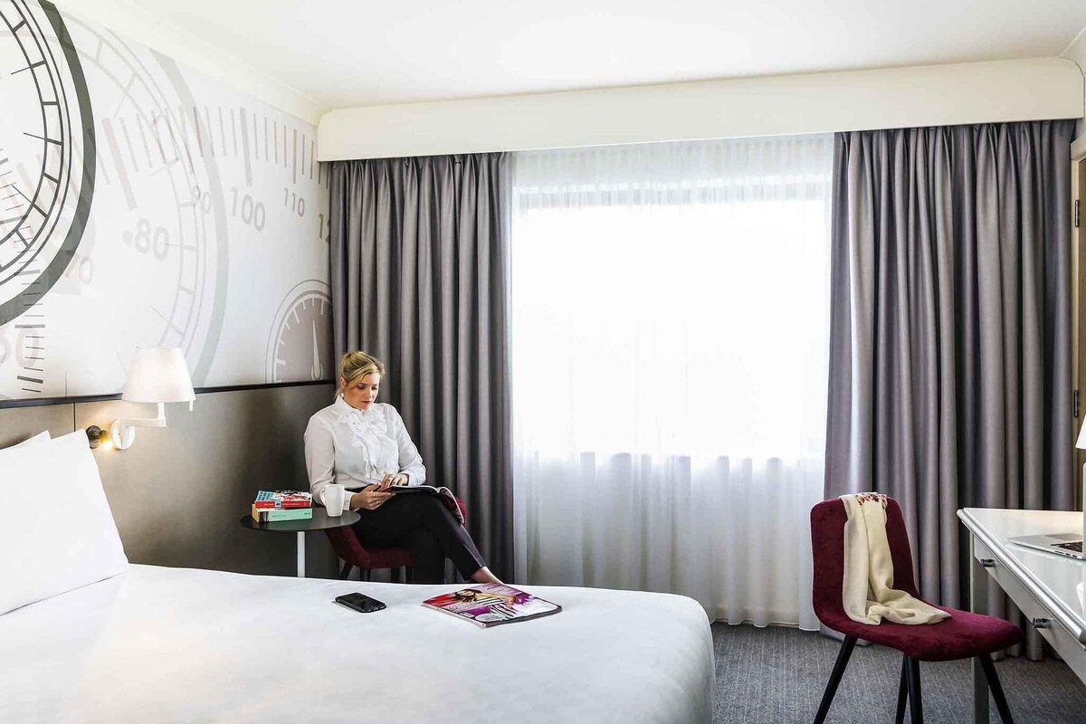 Una habitación de hotel con cama, escritorio, silla, lámpara y un reloj grande en la pared.
