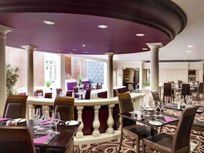 A hotel dining area with round tables, purple upholstery, and a view of the cityscape. - Mercure Dartford Brands Hatch Hotel & Spa (Sevenoaks)