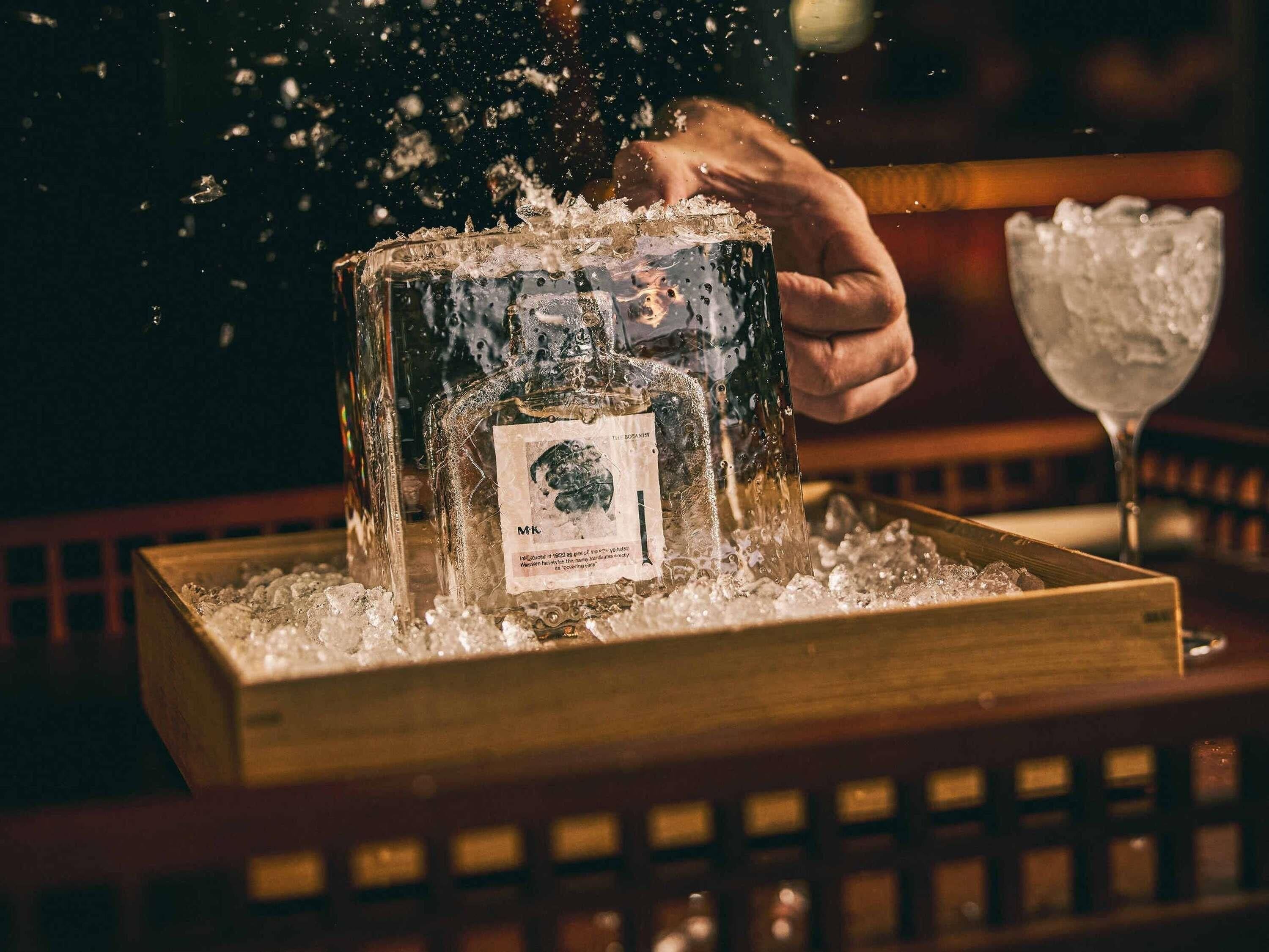Une bouteille de spiritueux posée sur de la glace, une main qui s’en saisit, et un verre d’eau glacée en arrière-plan.