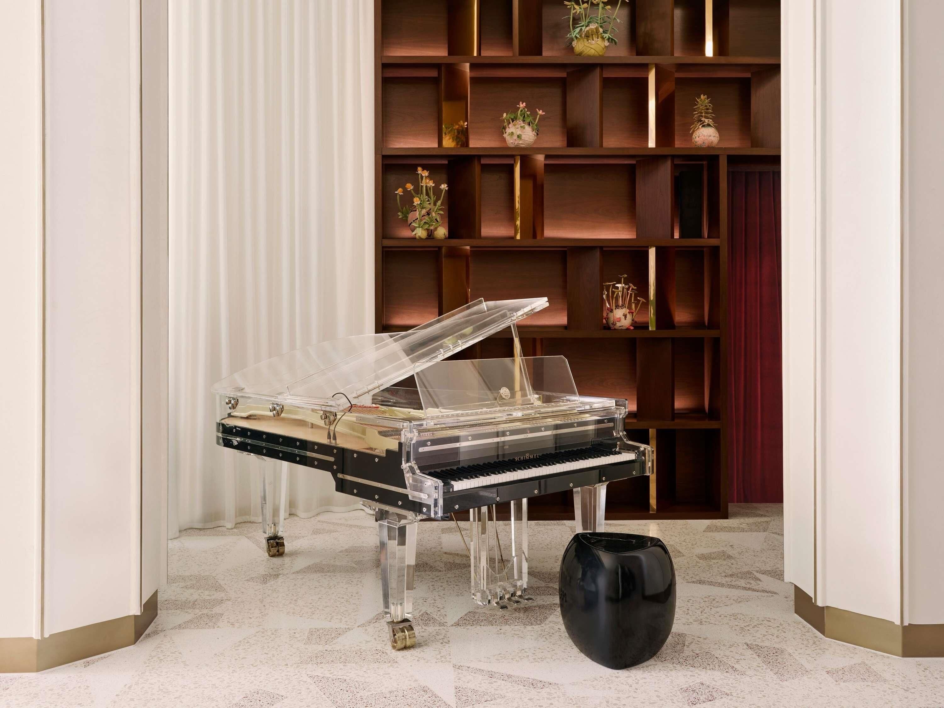 A grand piano in a corner with a clear protective cover, a black vase, and a wooden bookshelf with decorative items.