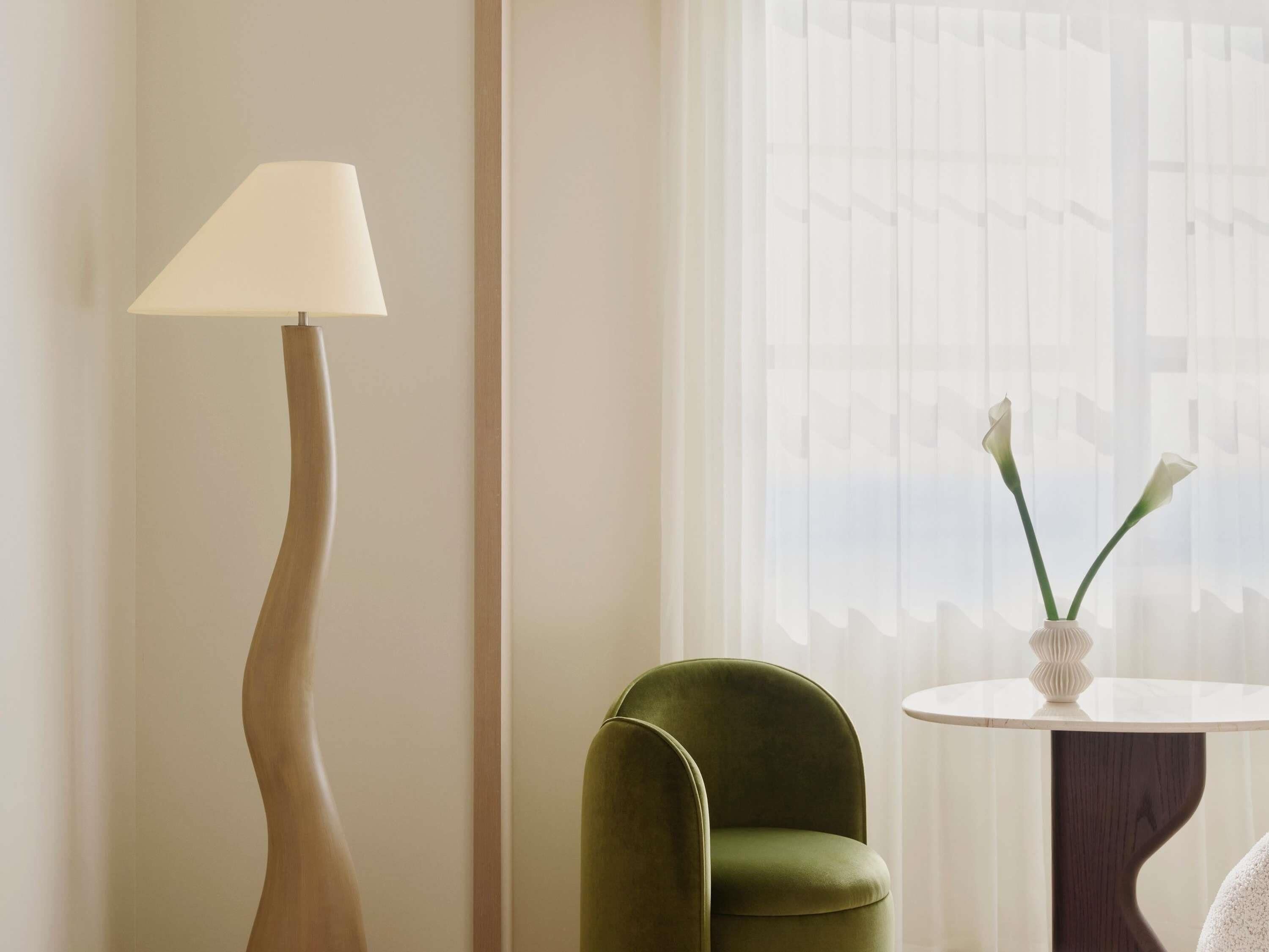 Una habitación con una lámpara de pie, un sillón verde, una mesa redonda con un florero y una ventana con cortinas de gasa.