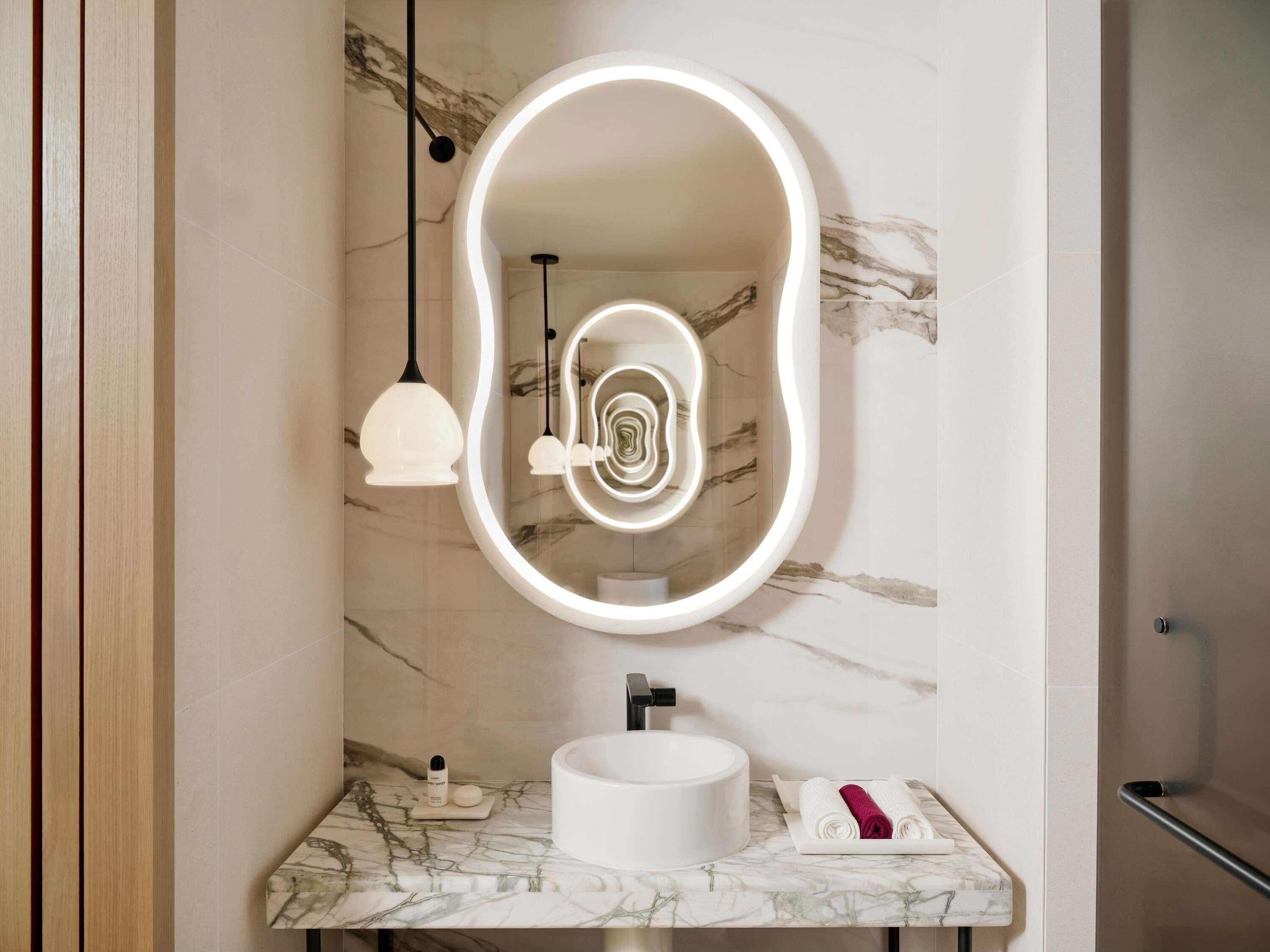 A bathroom with a marble sink, a large mirror with a light halo, pendant lights, and a towel rack.