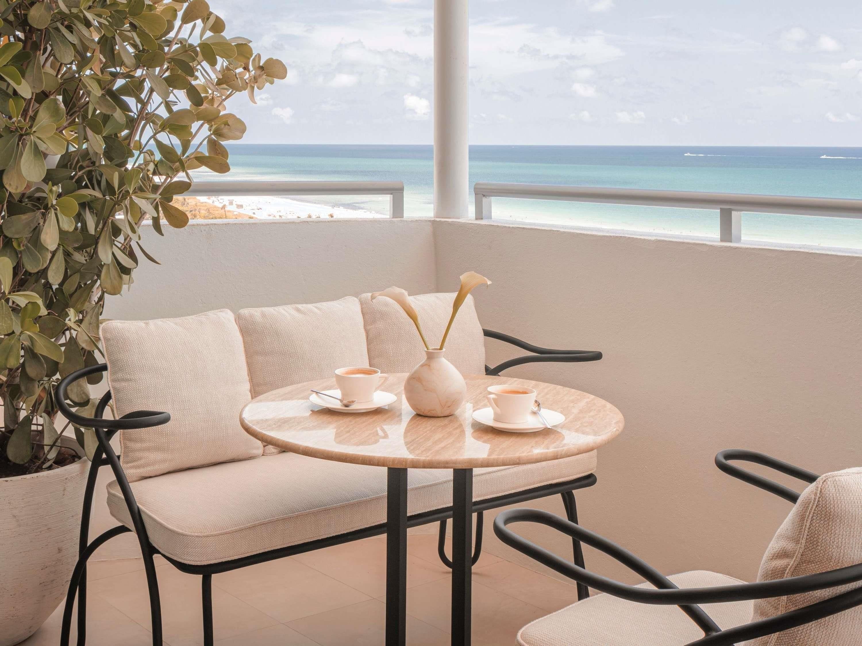 A balcony with a cushioned seating area, a round table with a vase and cups, and a view of the ocean.