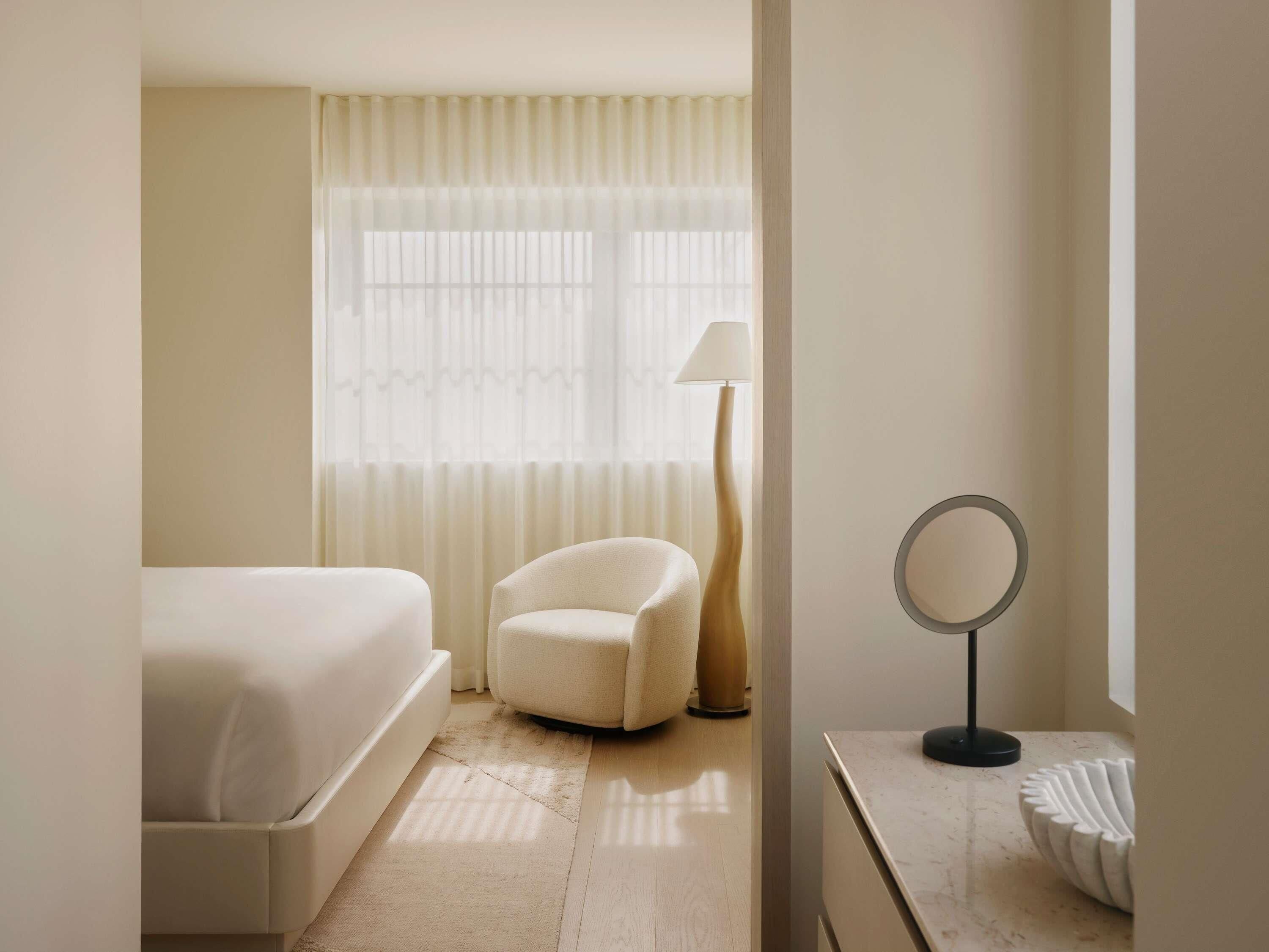 A hotel room with a bed, a chair, a desk with a lamp, and a mirror.