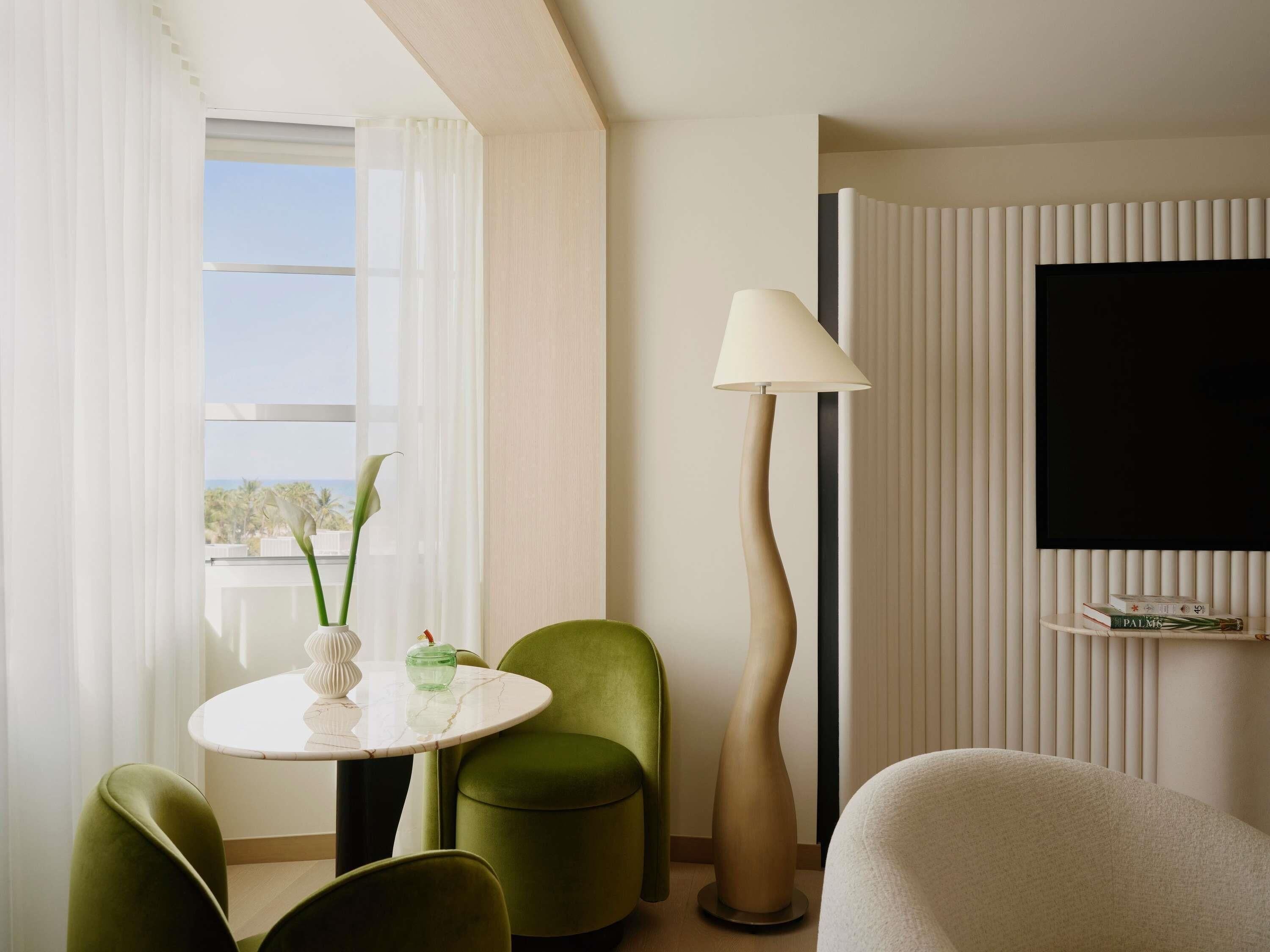 A modern hotel room with a round table, green chairs, a floor lamp, and a TV mounted on the wall.
