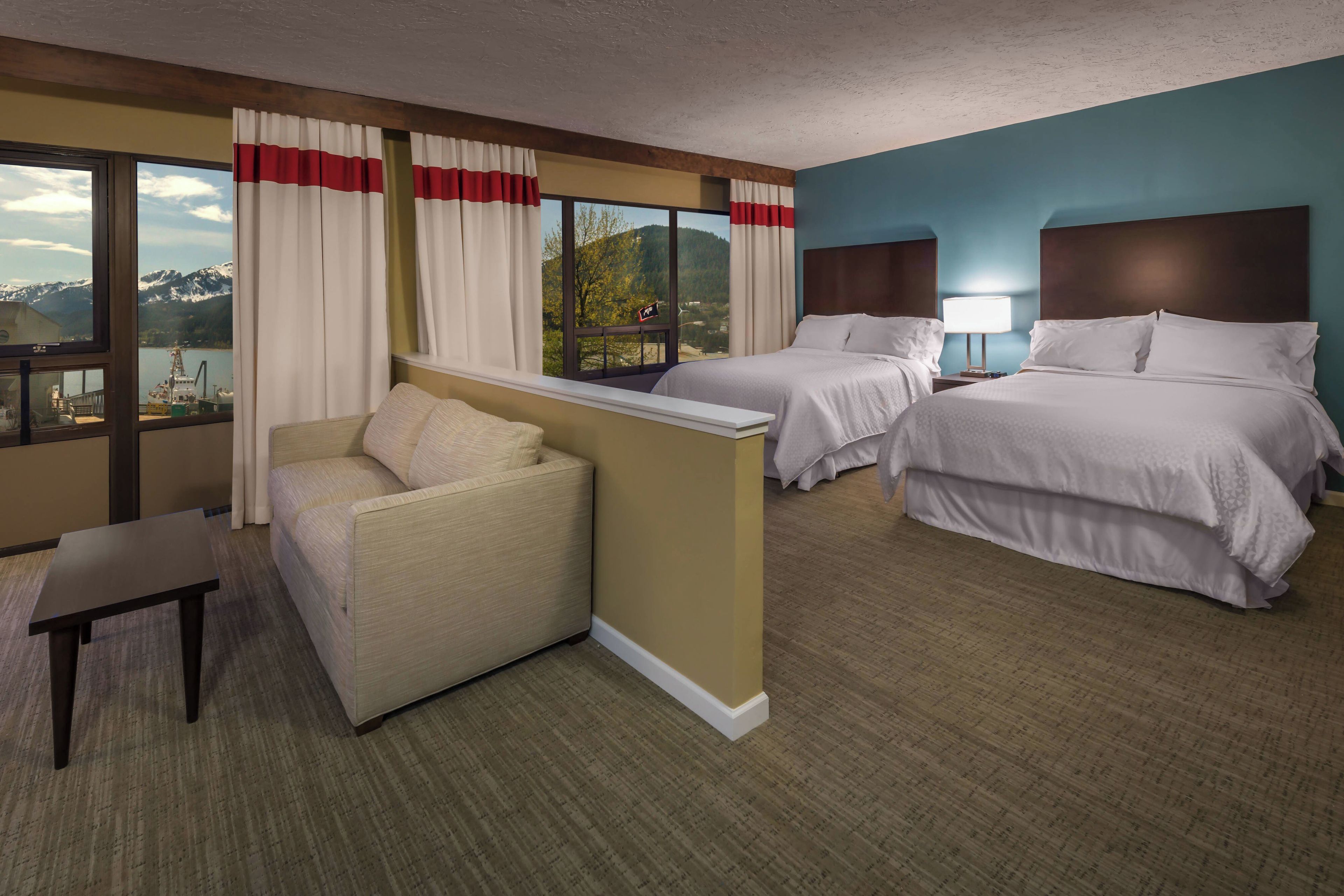 A hotel room with two beds, a sofa, a desk, and large windows with curtains.