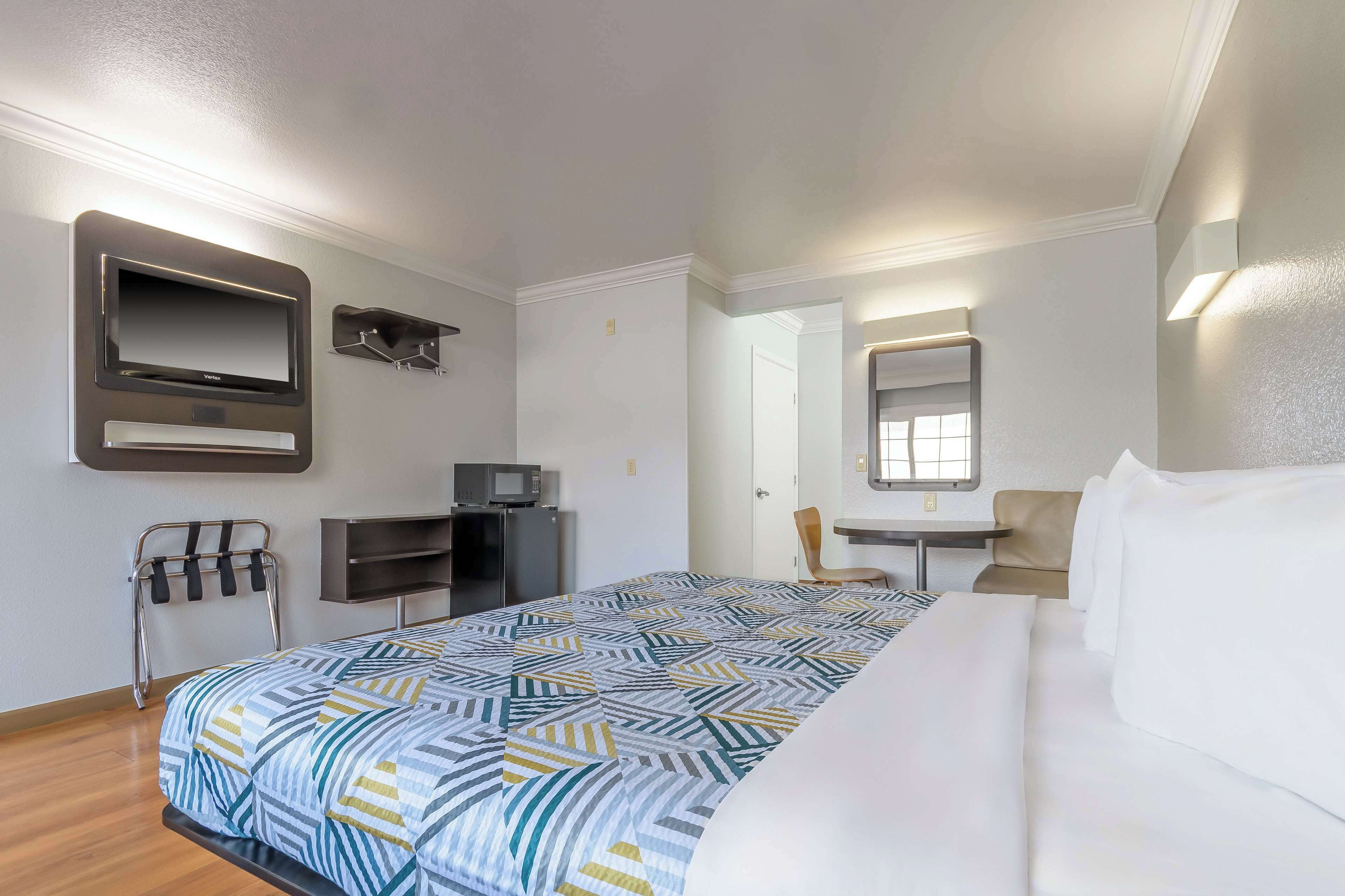 Quarto de hotel com uma cama, uma escrivaninha, uma cadeira, uma TV e um micro-ondas.
