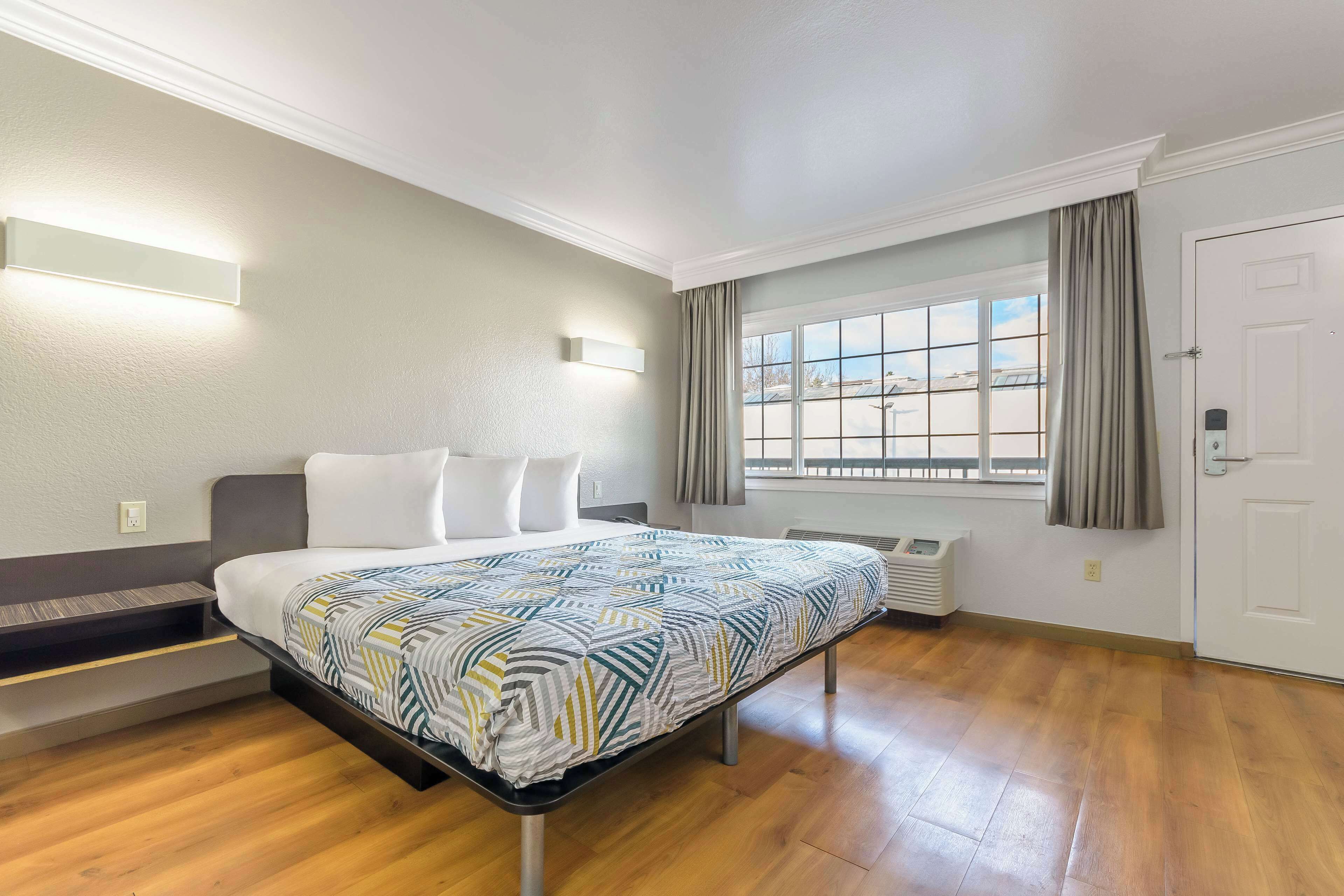 A hotel room with a bed, a window with curtains, a wall-mounted light, and a thermostat.