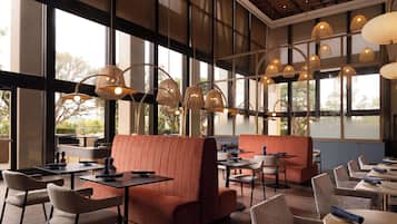Un restaurante moderno con techos altos, amplios ventanales y una variedad de opciones de asientos, incluyendo cabinas y mesas.