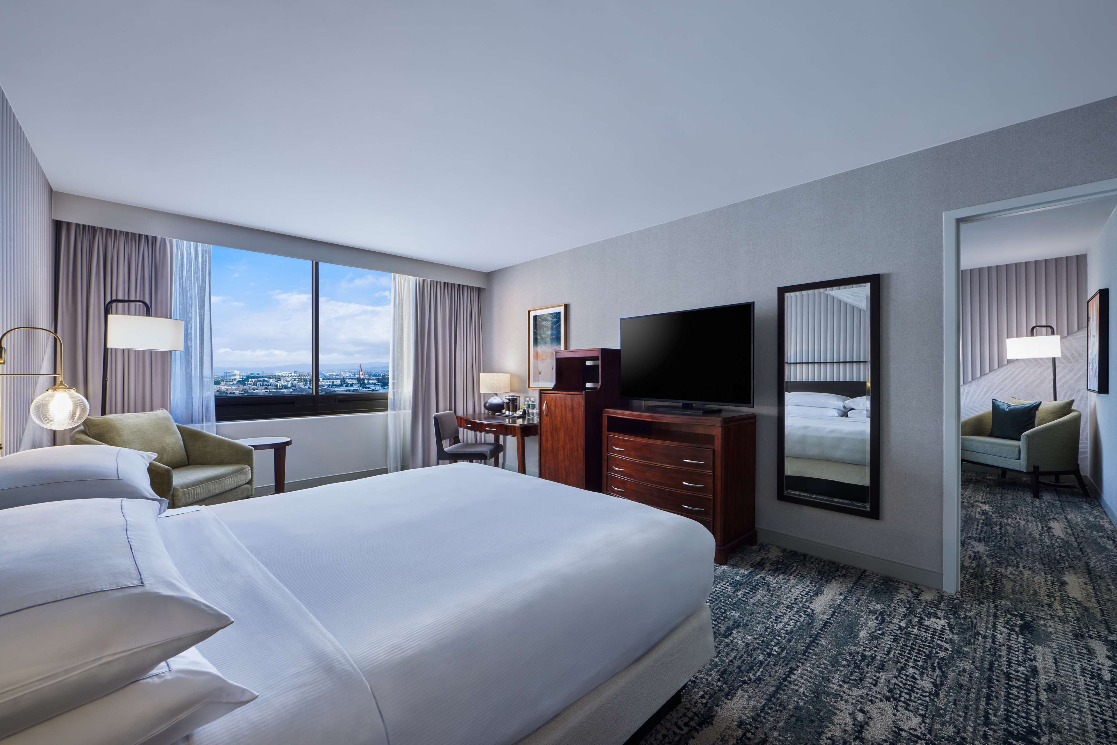 A hotel room with a large bed, a desk, a TV, and a view of the city.