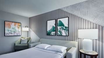 A modern hotel room with a bed, a chair, a lamp, and two framed pictures on the wall.